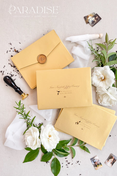 Soft gold wedding envelopes with printed addresses, styled flat lay with white florals, greenery, wax seal, and ribbon, perfect for elegant invitation suites.