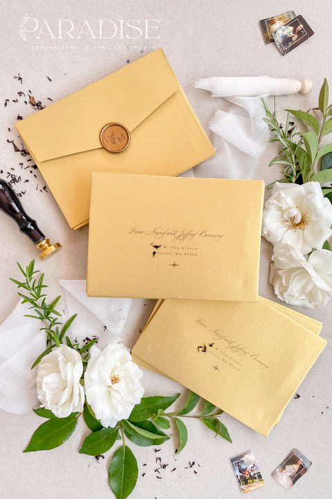 Soft gold wedding envelopes with printed addresses, styled flat lay with white florals, greenery, wax seal, and ribbon, perfect for elegant invitation suites.