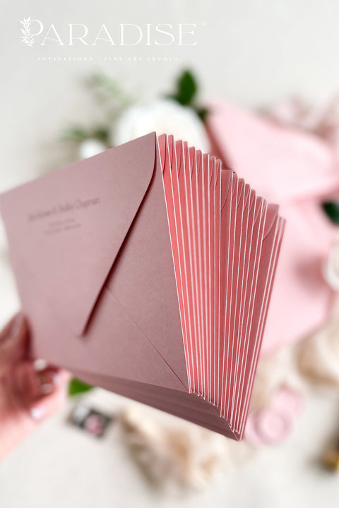 Dusty pink envelopes with custom address printing, displayed on a surface with a white rose and green leaves, indicating a possible use in wedding or formal invitation settings.