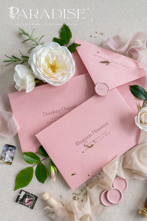 Dusty pink envelopes with custom address printing, displayed on a surface with a white rose and green leaves, indicating a possible use in wedding or formal invitation settings.