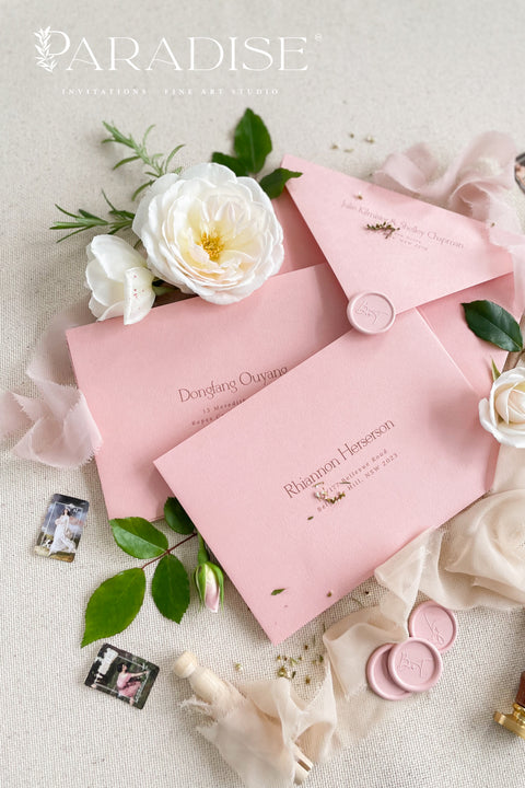 Dusty pink envelopes with custom address printing, displayed on a surface with a white rose and green leaves, indicating a possible use in wedding or formal invitation settings.